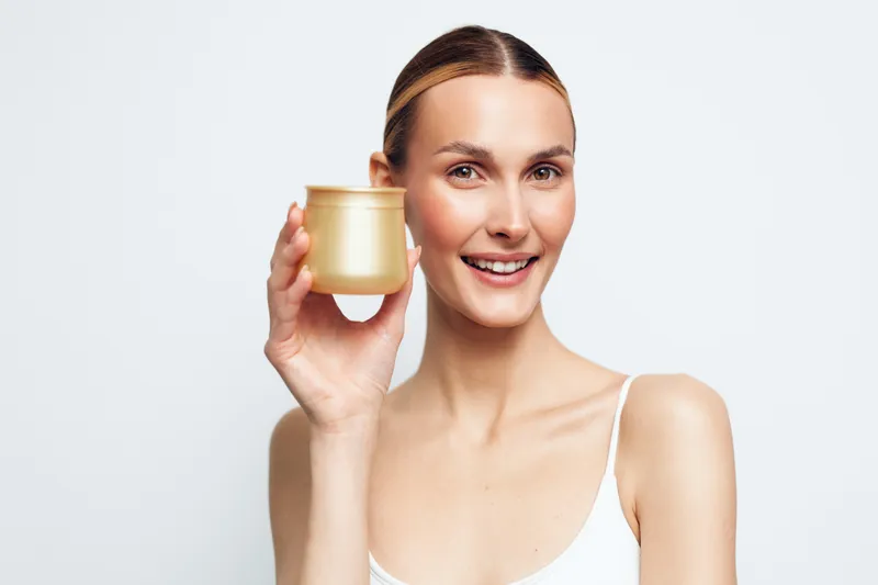 Woman skincare beauty moisturizer cream jar smiling portrait in studio. Young woman holds golden jar of facial cream, soft white background, skincare and wellness concept.