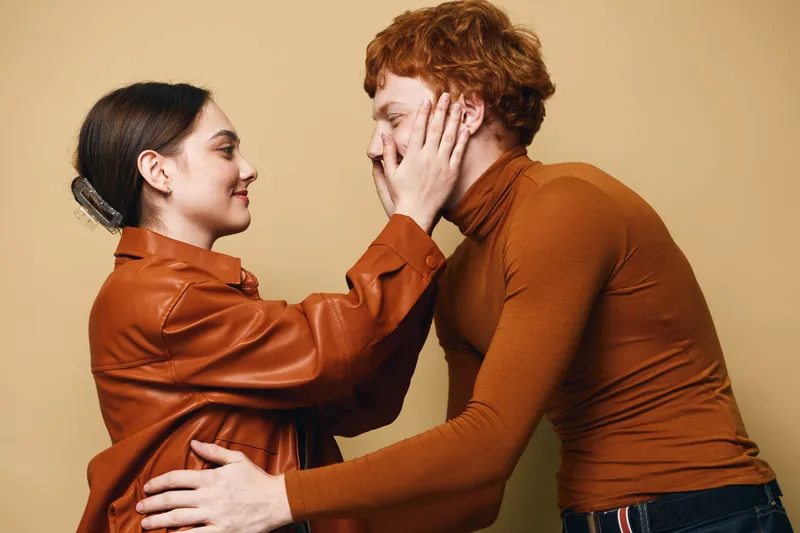 Young couple in casual clothing embracing and showing affection indoors. Woman holding man's face with hands, both smiling and enjoying tender moment together.