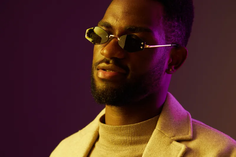 Stylish man wearing sunglasses and a beige coat against a purple background, exuding confidence and modern fashion trends