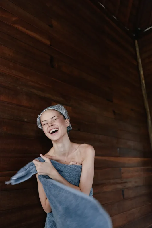 laughing woman wrapped in towel with headband enjoying wooden spa interior, natural light and joyful expression create warm relaxing atmosphere