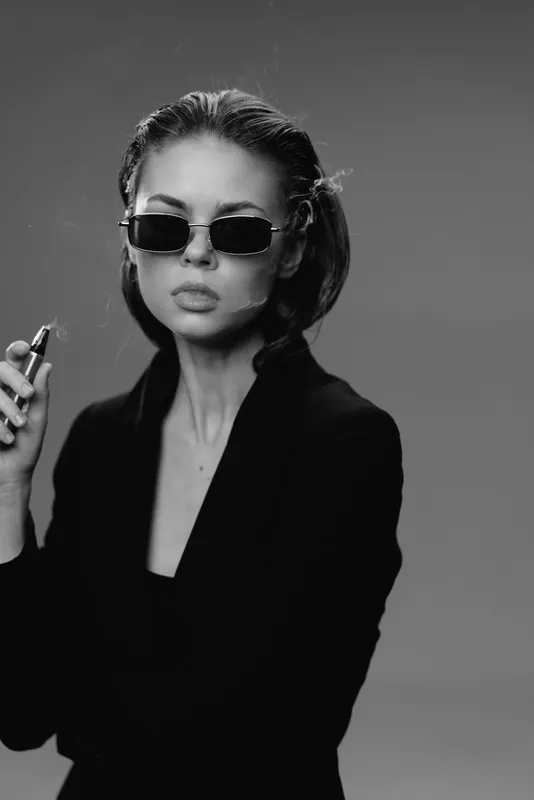 Black and white portrait of a stylish woman wearing sunglasses and holding lipstick, expressing confidence and elegance.