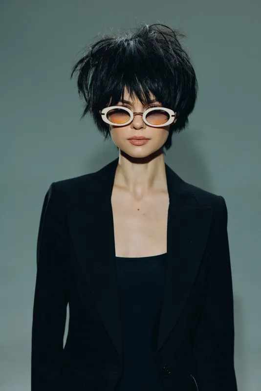 Stylish woman wearing oversized sunglasses and black blazer with short tousled hair and confident expression.