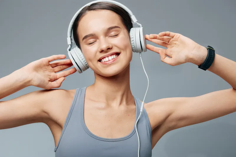 Fitness and sport themed image of a woman in a gray sleeveless top, wearing headphones, smiling with eyes closed, enjoying music during a workout in a clean studio setting.