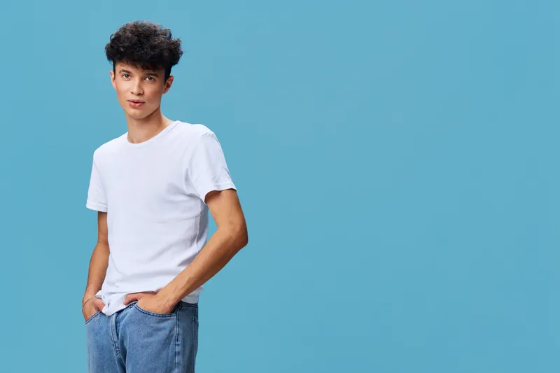 young man wearing casual white t-shirt and blue jeans standing confidently against blue background with hands in pockets, curly hair, relaxed posture
