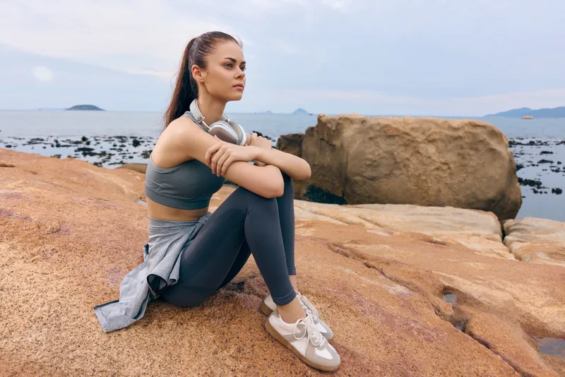 Athletic woman in activewear sitting on rocky beach, looking contemplatively at the sea, promoting fitness and wellness with a serene atmosphere