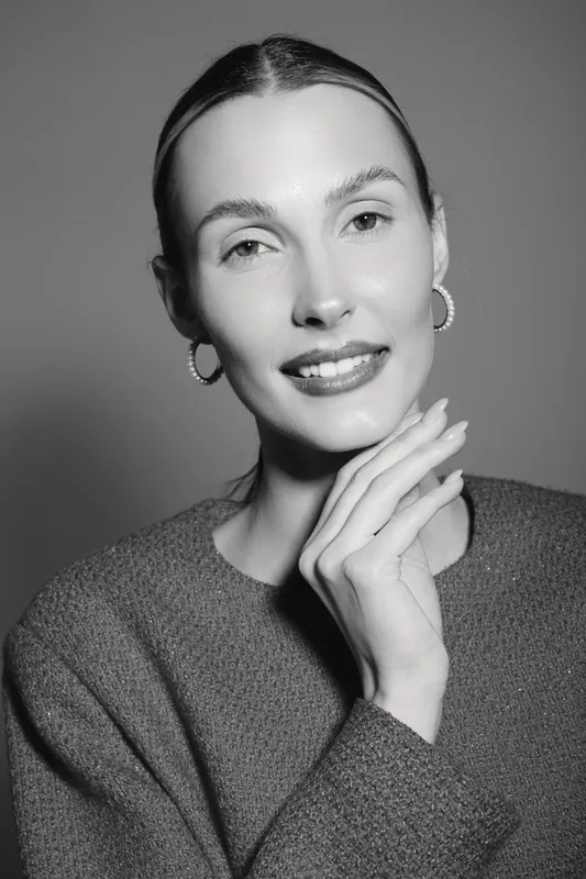 Smiling caucasian woman with fair skin and slicked hair poses with hand near face, wearing earrings and sweater in studio portrait. Concept of beauty and fashion.