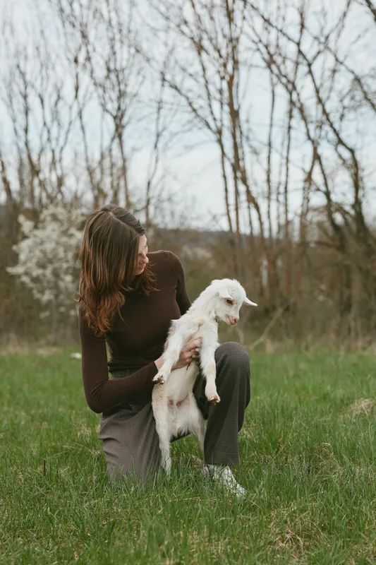 Young woman with long brunette hair kneels on grass holding a white baby goat in a natural outdoor setting during early spring with bare trees and soft light.