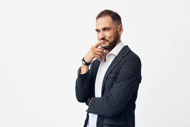 A professional businessman stands against a clean white background, exuding confidence and focus. He touches his lips with a thoughtful gaze, revealing calm determination and poised leadership.
