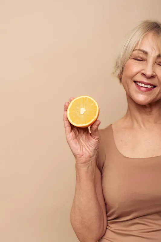 Smiling mature woman with short blonde hair holds half a fresh orange, eyes closed, wearing beige top on neutral background. Concept of healthy lifestyle, vitamin c, and happiness.