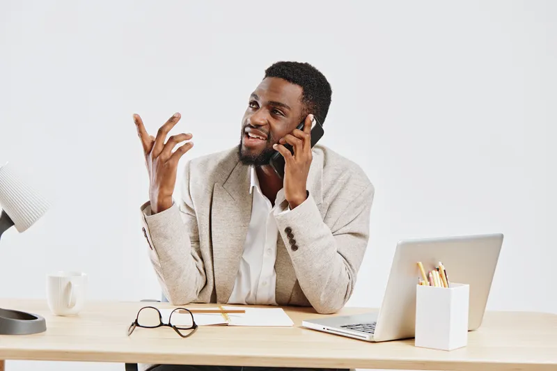 Smiling young African American man talking on smartphone while working at desk in modern office setting, using laptop for communication and multitasking, casual fashion, positive mood, studio shot, pr