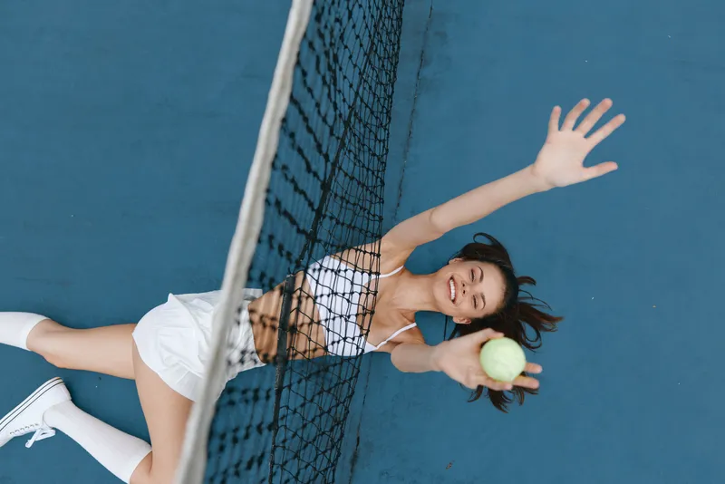 Happy young female tennis player in white sportswear reaching for a ball on a blue court, showcasing athleticism and joy in sport while emphasizing a vibrant and active lifestyle
