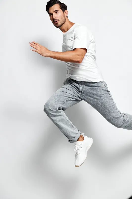 Active young man wearing a white t-shirt and gray joggers, energetically running against a plain light background. Concept: fitness and lifestyle motivation.
