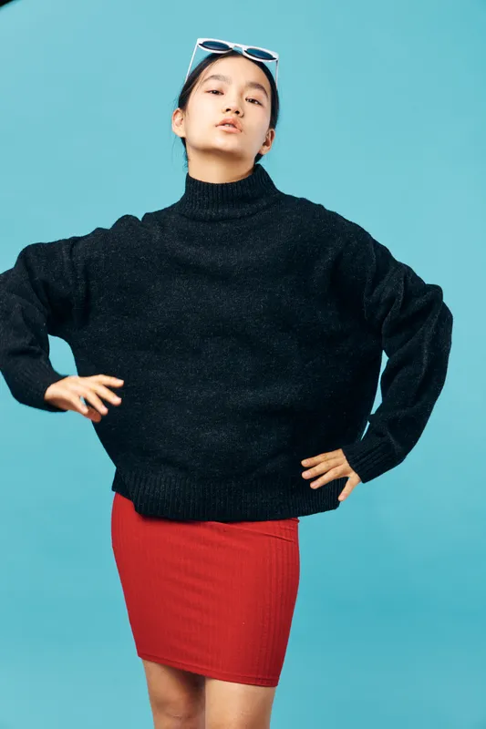 Young woman in black sweater and red skirt posing confidently with hands on hips against blue background, stylish casual fashion portrait with attitude and sunglasses on head.