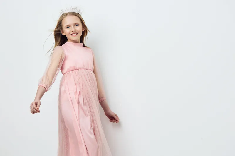 Cheerful young girl wearing a pink dress with a tiara, smiling joyfully against a minimalist white background, perfect for children s fashion and celebrations