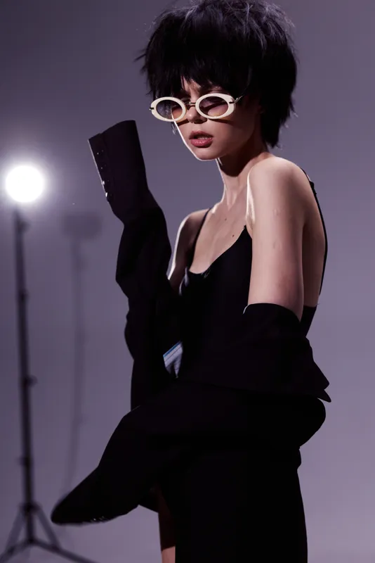 Stylish woman with short black hair wearing white round sunglasses and black clothing in a moody studio setting.