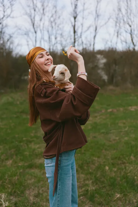 Smiling woman holding a small dog outdoors in a field with bare trees, wearing a brown coat and orange headband. Casual lifestyle moment capturing friendship, nature, and joy in soft natural light.