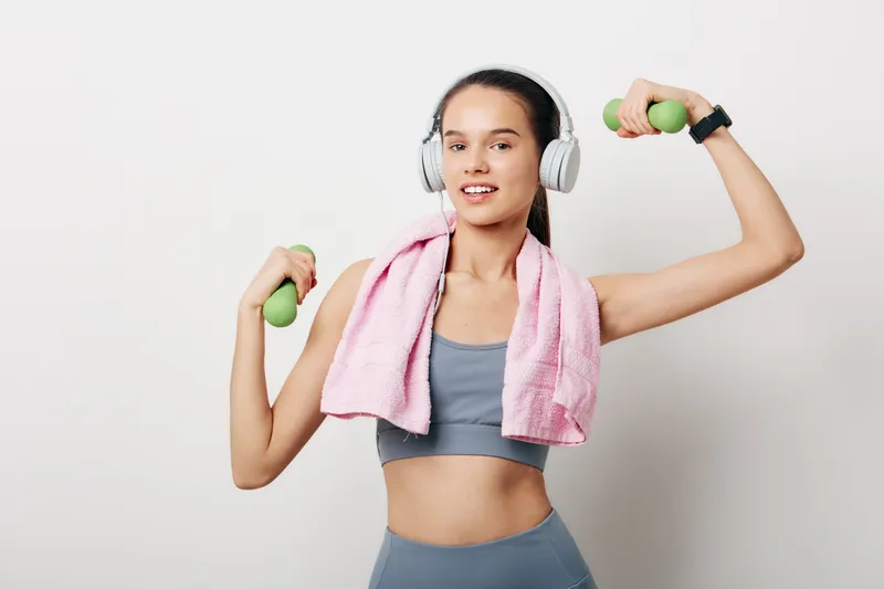 Dynamic portrait of a woman in workout gear exercising with dumbbells, headphones on, a pink towel around her shoulders, in a clean studio for fitness and sport inspiration.