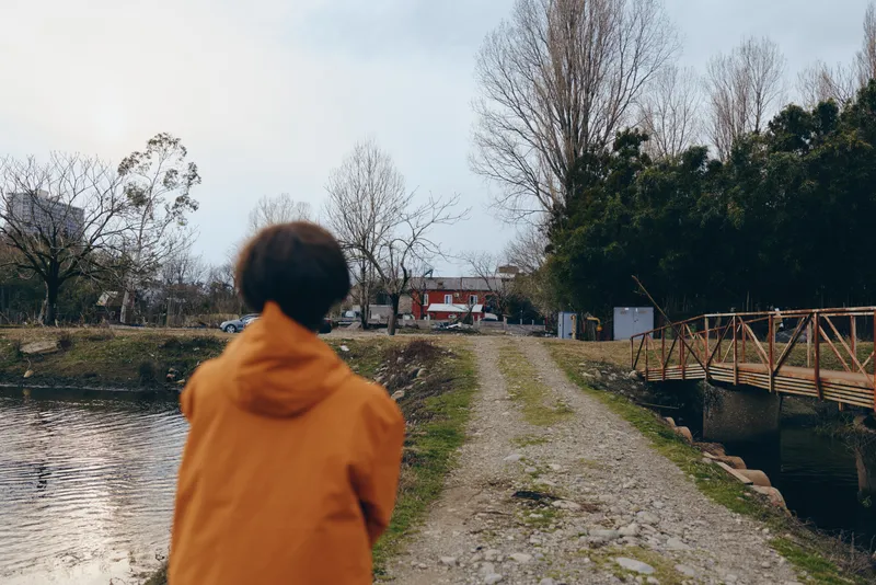 Woman lifestyle outdoors walking by river path, orange hooded jacket, back view to camera, cloudy sky, calm nature, bare trees, canal water reflection, slow stroll