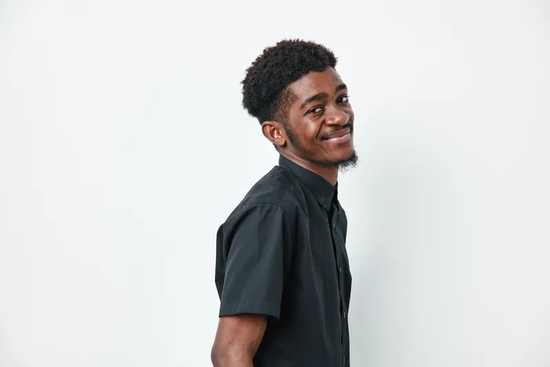 Young black man with dark skin and curly hair smiling and looking at camera wearing black shirt on white background in studio portrait.
