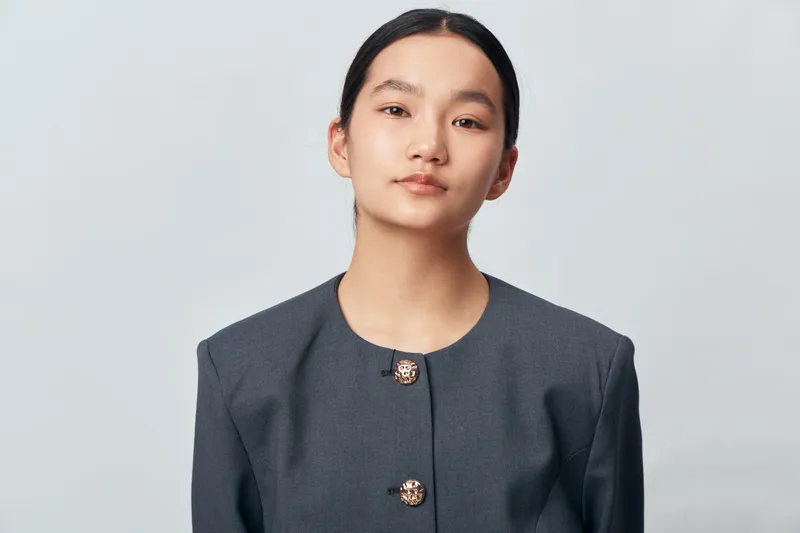 Portrait of young woman with natural makeup wearing grey jacket with buttons looking at camera against plain light background. Minimal style, confident expression and clean professional look.