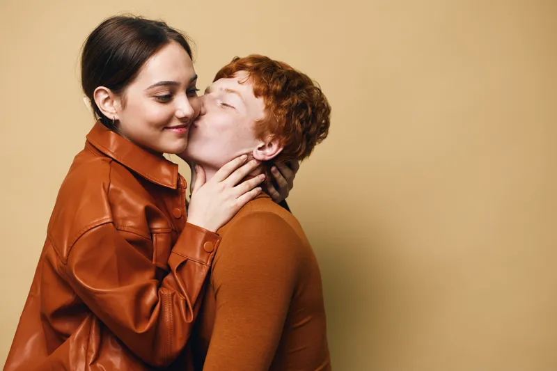 Couple, love, kiss and embrace with young woman and man in casual clothes on beige background. Romantic relationship, affection and happiness with ethnic diversity and intimacy closeup.