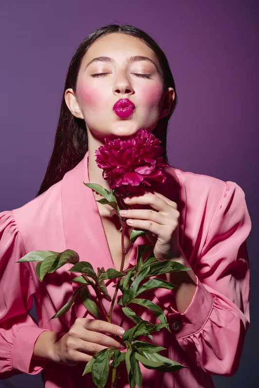 Beautiful young woman in a pink outfit with vibrant makeup kisses a bright flower while holding it gently. Studio lighting enhances the fashion and beauty concept creatively.