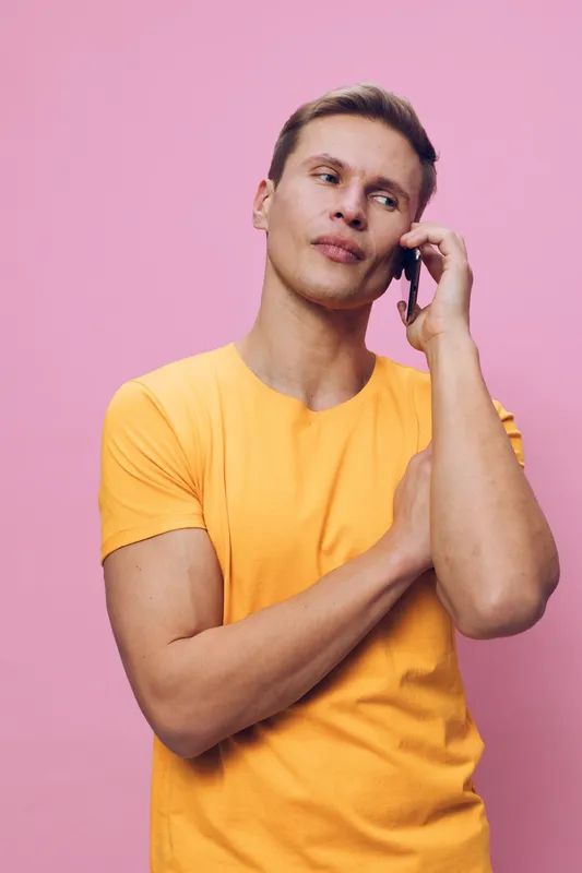 Confident young man wearing casual yellow t-shirt talking on mobile phone and looking thoughtful, standing against pink background, studio portrait. People lifestyle concept