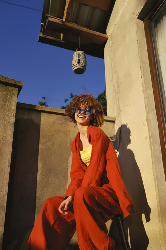 A stylish woman sits outdoors on a sunny terrace, wearing a red jumpsuit and sunglasses. A bold lifestyle portrait highlighting confidence, warmth, and carefree fashion
