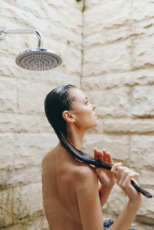 young woman showering outdoors under rainfall with wet hair and natural stone wall background, serene and refreshing moment in warm lighting