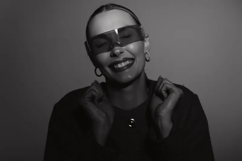 Smiling white woman with pale skin wearing futuristic visor and hoop earrings in studio with dark clothing and joyful expression, concept of modern fashion.