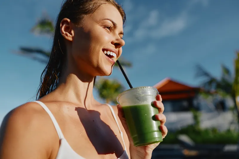 A healthy woman enjoys a green smoothie outdoors in bright sunlight, smiling as she sips through a straw. A lifestyle moment capturing refreshment, vitality, and wellness in a sunny, casual setting.
