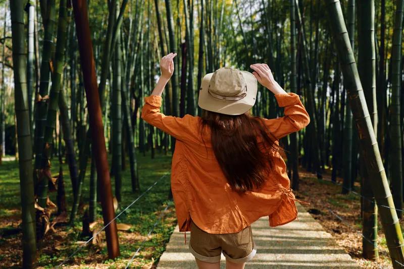 Woman bamboo forest hat path walking orange back view of traveler on a stone pathway, wearing shirt and shorts while exploring peaceful green grove in sunny outdoor summer scene