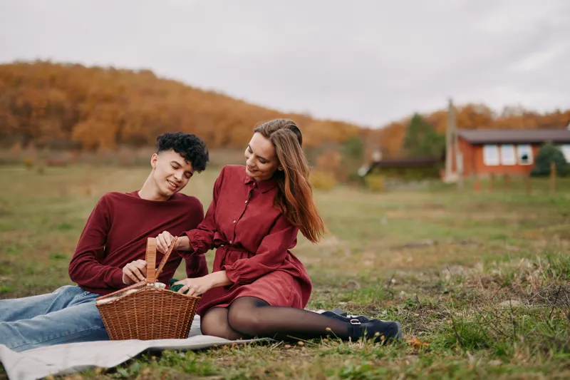 A genuine outdoor picnic moment showing an authentic couple sharing food from a wicker basket in a natural autumn field, capturing sincere connection and everyday warmth.