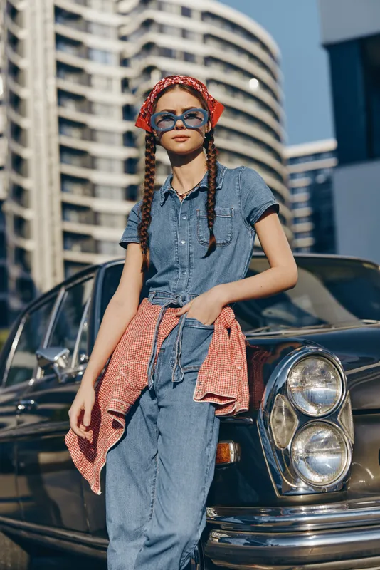 Lifestyle woman in modern boho-western denim street style with braided hair and retro sunglasses standing confidently by a vintage car in film color tones.