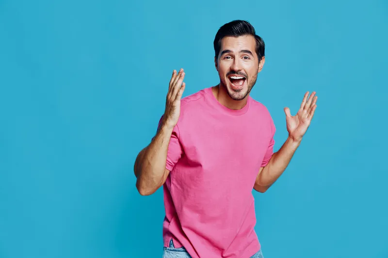 Happy cheerful young man wearing pink sweater expresses excitement with raised hands against a bright blue background. People lifestyle concept.