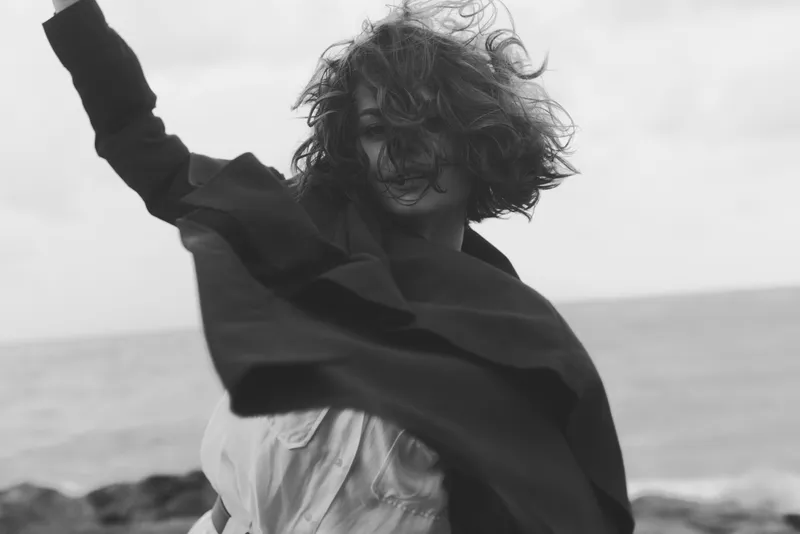 Windy outdoor scene with scarf and coat in motion by the ocean, hair swept by breeze and monochrome tones, capturing dynamic movement