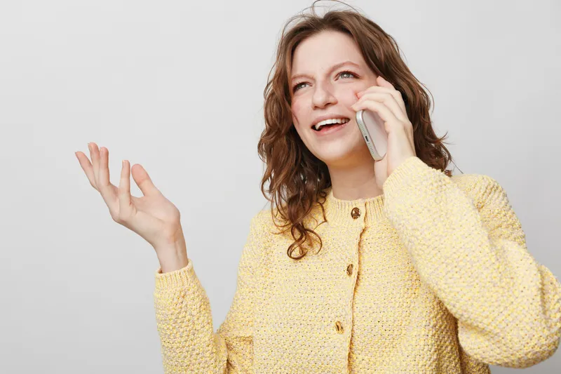 Smiling woman with curly hair wearing yellow sweater talks on mobile phone, gesturing with hand. Bright studio background. Concept of communication, happiness, and casual lifestyle.