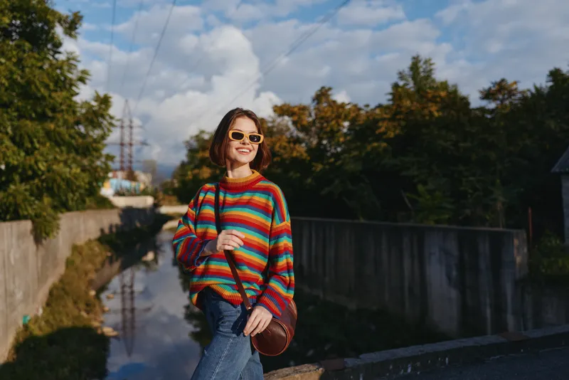 Young woman in her mid twenties wearing a colorful striped sweater and sunglasses, smiling while walking on an urban canal bridge with crossbody bag and casual jeans, lifestyle travel.