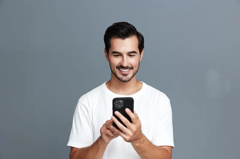 Smiling man wearing a white t-shirt looks at mobile phone with joy, standing against a plain gray background. Modern lifestyle and technology concept.