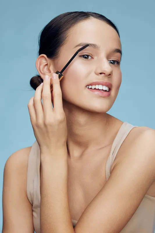 Young woman applying eyebrow gel in light natural makeup, showcasing beauty and confidence with a soft blue background, perfect for beauty and skincare themes
