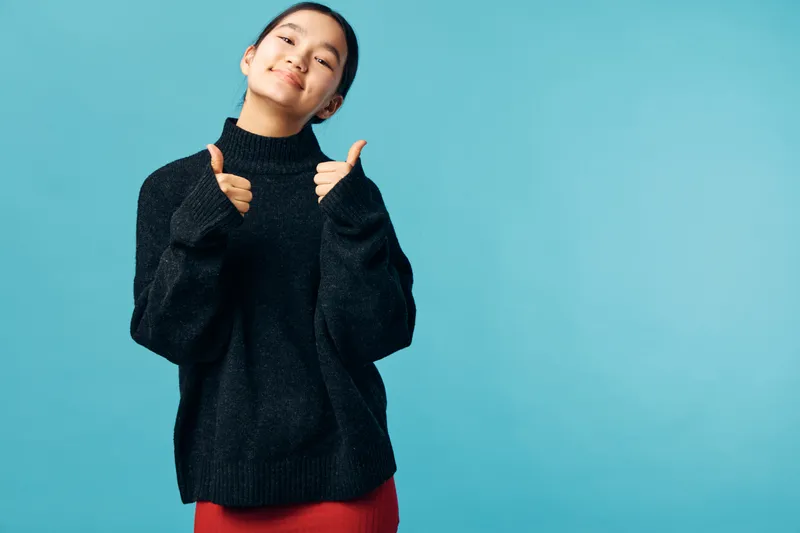 Young woman standing in front of blue wall showing thumbs up and smiling, wearing black oversized sweater and red skirt with casual expression and positive mood.