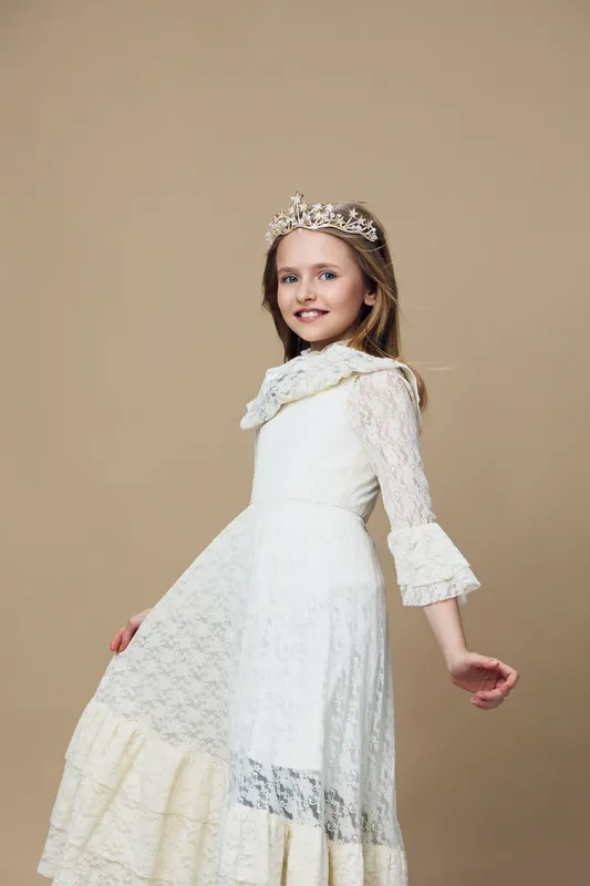 Elegant young girl in a white lace dress with a crown, exuding joy and confidence against a neutral background, perfect for family portraits or celebrations