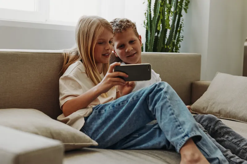 Two children sitting on a sofa looking at a smartphone screen together with smiles. Casual home setting with natural light and green plants in the background, concept of family and technology.
