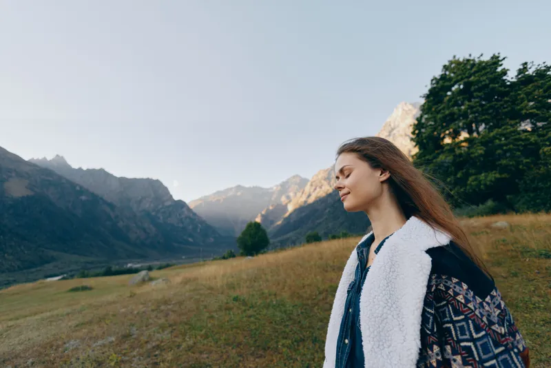 Woman stands in meadow near mountains, portrait shot outdoors during sunrise, travel lifestyle scene showing peaceful landscape, young female in warm coat enjoying nature.
