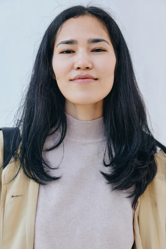Woman with long dark hair wearing a beige coat and knit sweater stands outdoors with a calm expression, soft natural light, minimal fashion style and everyday chic, clean background, in frame