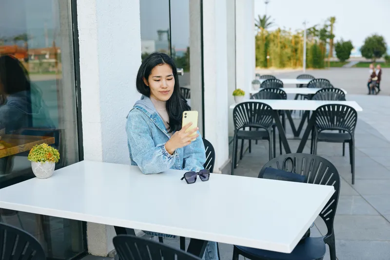 Woman seated at outdoor cafe using smartphone, denim jacket, sunglasses on white table, enjoying a calm moment in sunny weather during a casual day