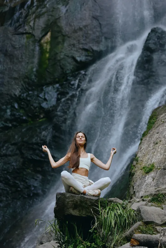 Woman meditating in white clothes seated on rock in front of a large waterfall, surrounded by lush greenery and natural scenic beauty during daylight.