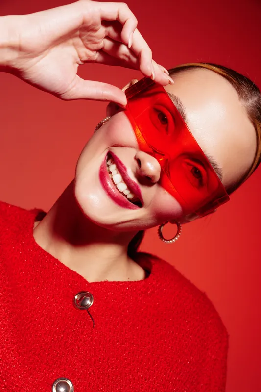 Smiling woman in red sweater and red transparent sunglasses poses against red background. Fashion portrait with bright colors and joyful mood, concept of style and confidence.