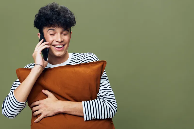 smiling man talking phone holding pillow with closed eyes wearing striped shirt, curly hair, relaxed mood on green background, indoor casual lifestyle