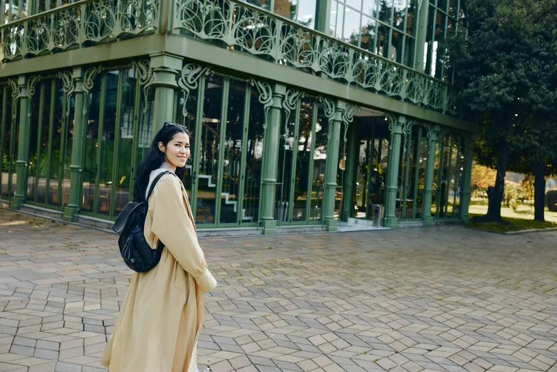 Beige coat, backpack, and traveler pose beside a glass greenhouse in a park, capturing curiosity, fashion, and outdoor exploration under soft daylight